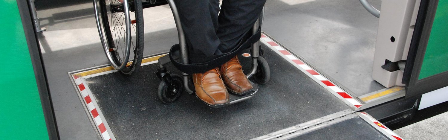 Das Bild zeigt die Beine eines Rollstuhlfahrers, der über eine ausklappbare Rampe aus einem öffentlichen Verkehrsmittel fährt.